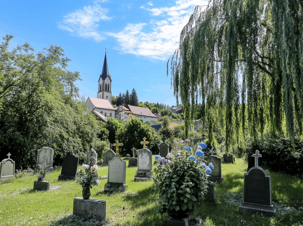 Grüner Friedhof mit Grabsteinen und Blumen, im Hintergrund Kirche - würdevolle Bestattung in Bayern.